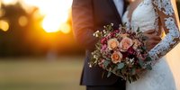 Romantic wedding couple holding peach and burgundy rose bouquet with lace sleeve detail against golden sunset background, intimate outdoor ceremony moment.