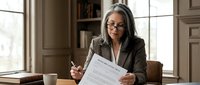 Mature woman with gray hair and glasses wearing business attire reviewing documents at desk in modern office setting, suitable for corporate, professional, and business concepts.