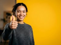 Young woman with dark curly hair and medium brown skin tone giving thumbs up gesture against yellow background, expressing approval and positivity for success and motivation concepts.