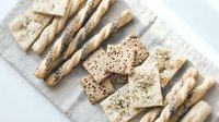 Assorted whole grain and seeded crackers and breadsticks arranged on linen cloth, showcasing variety of healthy snack options for food styling and culinary projects.