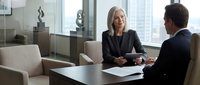 Senior woman executive in dark blazer conducting business meeting with male colleague in modern office conference room with city skyline view.