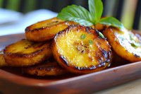 Caramelized plantain slices garnished with fresh basil leaves on wooden plate, golden brown crispy edges and soft interior, close up view of tropical dessert.