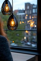 Modern pendant lights with glowing filament bulbs in smoke glass shades against city window view at dusk, creating cozy atmospheric lighting and bokeh effect.