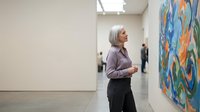 Middle-aged Caucasian woman with gray bob haircut viewing colorful abstract artwork in modern white gallery space with visitors in background.