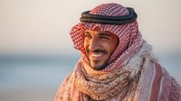 Middle Eastern man in traditional keffiyeh and scarf smiling warmly at sunset, with soft coastal background creating peaceful atmosphere.