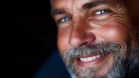 Confident mature Caucasian man with gray beard and bright smile against dark background showing genuine happiness and positive aging concept.