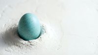 Single turquoise speckled egg nestled in white flour on clean surface for Easter baking and spring cooking projects.