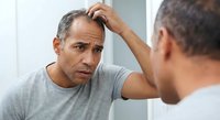 Middle-aged Hispanic man examining hair loss in bathroom mirror, touching scalp with concerned expression. Male pattern baldness, thinning hair, grooming routine.