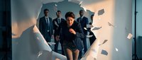 Young businesswoman breaking through paper wall with male colleagues in background representing breakthrough success and leadership in corporate environment.