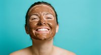 Young woman with chocolate face mask smiling joyfully during spa treatment on turquoise background for skincare and wellness routine.