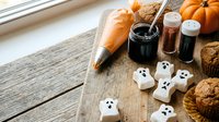 Halloween baking scene with ghost marshmallows, pumpkins, spices and muffins on rustic wooden table for seasonal cooking and party preparation.