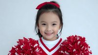 Young Asian girl with cheerful smile wearing red and white cheerleader outfit with pom poms against light background.