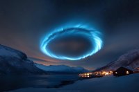 Mysterious glowing blue portal ring in night sky above snowy mountain landscape with illuminated village and fjord, creating magical atmospheric scene.