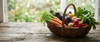 Fresh organic vegetables in wicker basket on wooden table by window, including carrots, tomatoes, eggplant and leafy greens for cooking and healthy eating.