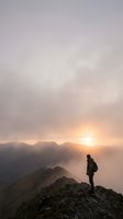 Hiker standing on rocky mountain peak at sunrise, gazing over misty valleys and distant ridges. Male adventurer wearing backpack, contemplating expansive landscape during golden hour. Suitable for travel, adventure, motivation, and outdoor lifestyle content.