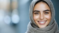 Young woman wearing grey hijab smiling warmly at camera with natural makeup and bright white teeth against blurred background. For diversity and inclusion content.