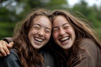 Two young Asian women with long brown hair laughing together outdoors, showing genuine joy and friendship while embracing. Natural lighting and green background create warm atmosphere.