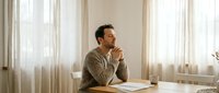 Young professional man in casual sweater sitting at wooden desk in bright office space, looking thoughtfully out window while working on laptop computer.
