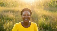 Young African woman wearing bright yellow shirt smiling at camera in golden hour sunlight among tall grass field, expressing joy and positivity for lifestyle and wellness content.