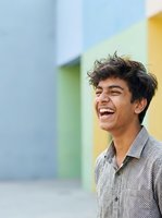 Young man with dark hair and South Asian ethnicity wearing light checkered shirt, laughing outdoors against colorful modern building backdrop. Suitable for lifestyle, happiness, youth culture.