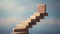 Wooden blocks stacked in ascending staircase pattern with target bullseye symbol on top block against blurred background, representing business growth, success achievement, and strategic goal progression.