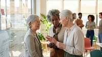 Three mature women of diverse ethnicities engage in conversation at a modern office workspace with large windows, potted plants, and architectural plans visible in the background, suggesting a collaborative design or planning meeting environment.