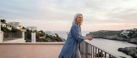 Mature blonde woman in blue dress enjoying sunset on Mediterranean terrace overlooking coastal village and sea. Relaxation and luxury lifestyle concept.