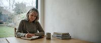 Mature woman with gray hair, Caucasian ethnicity, reading a book at wooden desk by window with coffee cup and stacked books nearby, indoor study environment.