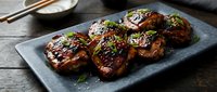 Grilled chicken thighs with teriyaki glaze and sesame seeds on slate plate. Asian cuisine dish garnished with green onions for restaurant menu design.