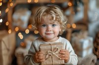Smiling toddler with curly blonde hair holding wrapped gift box against blurred Christmas lights background, wearing cozy beige sweater, festive holiday mood.