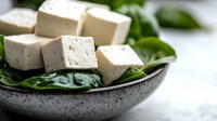 Fresh cubed tofu served on green spinach leaves in rustic ceramic bowl against light background, healthy vegetarian protein source for cooking.