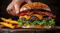 Juicy double cheeseburger with melted cheese, crispy bacon, fresh lettuce, tomato on sesame bun served with golden french fries on rustic wooden board in dark setting.
