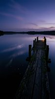 Wooden dock extending into calm lake at twilight with lantern glowing, surrounded by purple and blue sky, forested shoreline, and still water reflections for travel and nature projects.