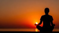 Man silhouette meditating in lotus pose at sunset over ocean. Peaceful yoga practice with warm orange sky background for wellness and mindfulness concepts.