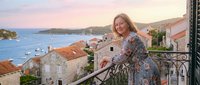 Mature blonde woman in floral dress enjoying sunset on Mediterranean balcony overlooking coastal town with terracotta roofs and azure sea waters.