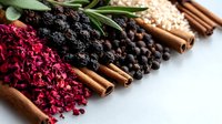 Aromatic spices and dried ingredients arranged in rows including cinnamon sticks, black peppercorns, dried red flowers, and herbs on white surface for culinary use.