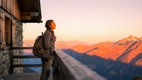 Middle-aged male hiker with backpack standing on mountain cabin balcony at sunrise, gazing at alpine peaks and valleys below during outdoor adventure and travel exploration.