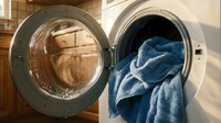 Modern washing machine with open door and blue towels spilling out in bright laundry room. Household chores and domestic cleaning concept for home care.
