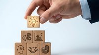 Businessman hand placing wooden cube with lightbulb icon on stack of business symbol blocks representing innovation and strategic planning concepts.