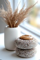 Chocolate frosted donuts with coconut sprinkles stacked near white ceramic vase with dried pampas grass, soft window light creates cozy minimal composition.