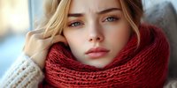 Young woman wearing cozy red knitted scarf and white sweater looking thoughtfully at camera with natural makeup and blue eyes on blurred background.