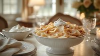Fresh homemade banana pudding topped with whipped cream and vanilla wafers in white ceramic bowl, served on elegant dining table with soft natural lighting.