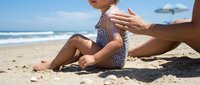Parent applying sunscreen to young child's arm on sandy beach with ocean and blue sky in background, sun protection and family skincare concept.