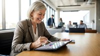 Mature blonde businesswoman using tablet computer in modern office environment with colleagues working in background during daytime meeting.