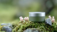 Minimalist metal cosmetic jar on green moss with pink flowers against blurred natural background, organic skincare product photography styling.