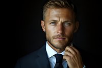 Confident Caucasian businessman in dark suit and tie against black background, thoughtful expression with hand near face, dramatic studio lighting creates professional mood.