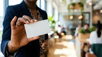 Male professional with dark skin tone wearing navy blazer holding blank white business card in modern office environment with blurred background and natural lighting.