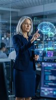 Mature woman with white hair, Caucasian ethnicity, wearing dark blue business suit, presenting holographic brain visualization in modern tech office environment for artificial intelligence and innovation concepts.