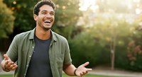 Young man with dark hair and olive complexion wearing casual olive shirt over dark t-shirt, smiling outdoors in natural sunlight with blurred green foliage background. Suitable for lifestyle, happiness, and casual fashion concepts.