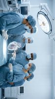 Surgical team of diverse medical professionals in sterile blue uniforms and caps performing operation in modern hospital operating room with advanced equipment and overhead lighting.